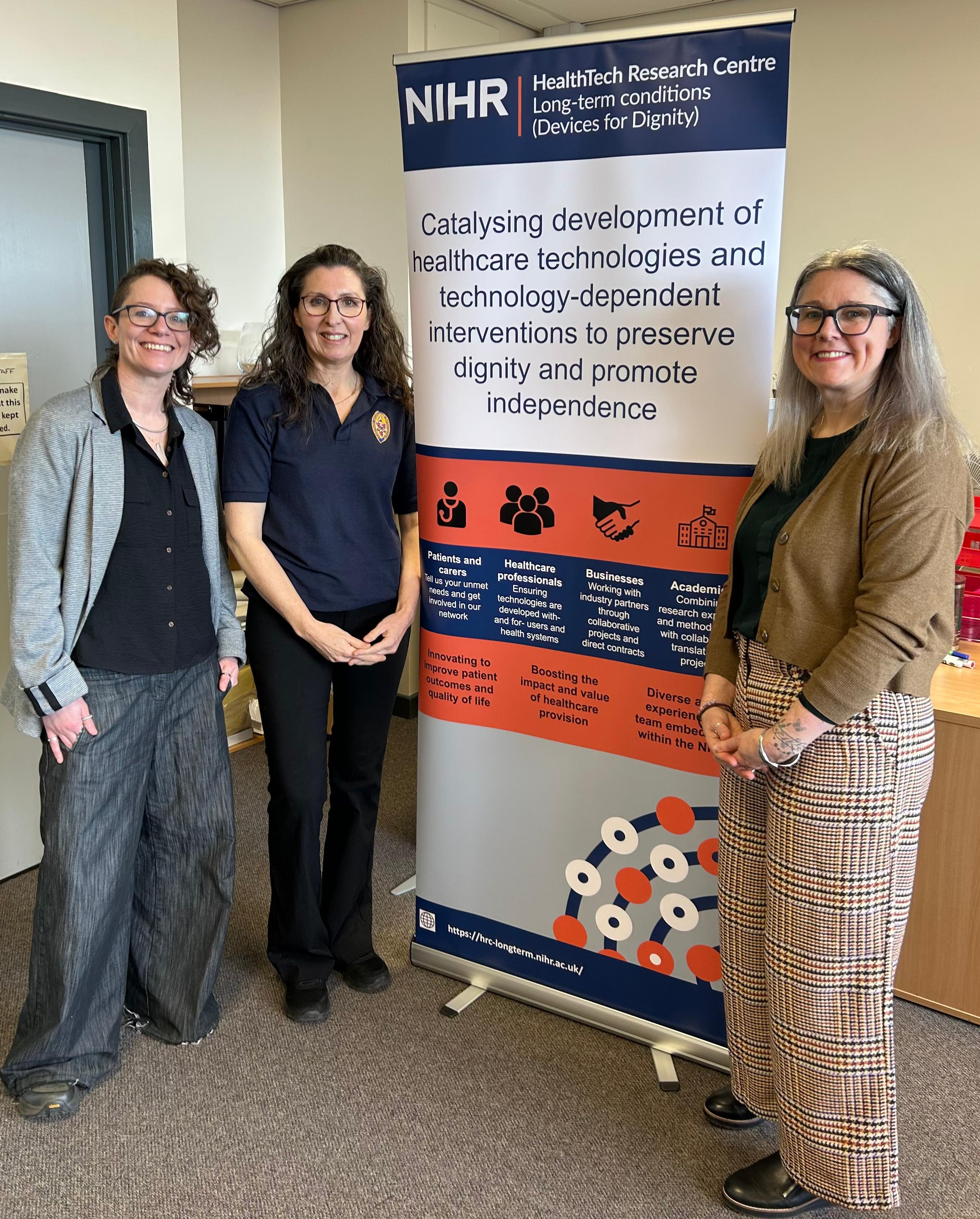 From left to right: Hayley Morris, Deputy Head of Operations: NIHR HRC in Long term conditions (Devices for Dignity), Jennifer Turner: JT Rehab, Liz Pryde, Head of Operations: NIHR HRC in Long term conditions (Devices for Dignity).  They are smiling at the camera and standing next to an NIHR-branded roller banner that details the work of the HRC.