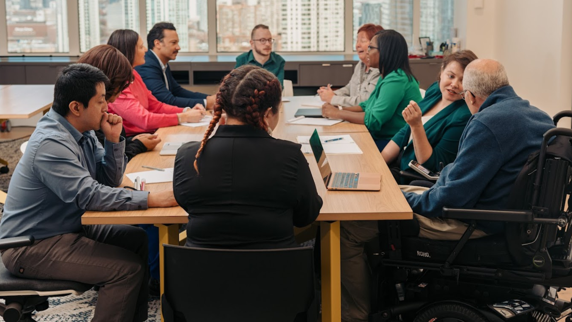 A diverse group of coworkers sit around a conference table in a modern office, engaged in discussion. One person uses a wheelchair, and a laptop and notebooks are on the table, with large windows and a cityscape visible in the background.