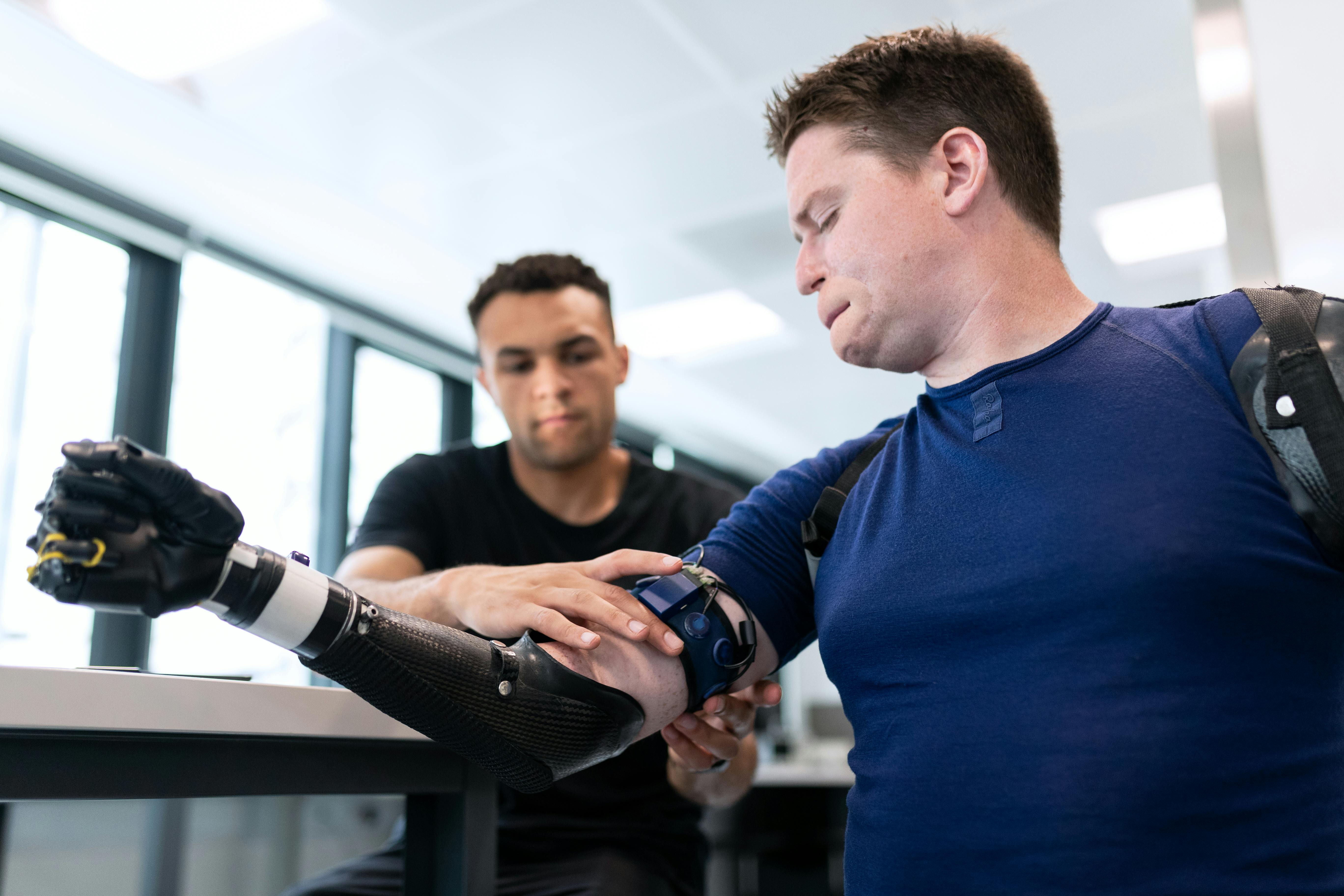 A man wearing a prosthetic forearm rests it on a table while another person adjusts a device attached near his elbow in a bright clinical or rehabilitation setting.